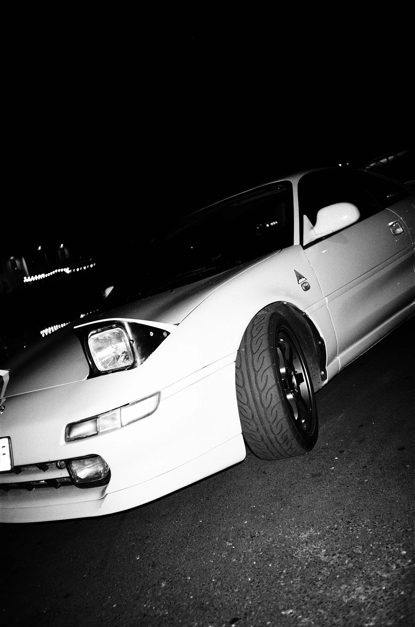 A black and white photo of a parked car at night, showing the front and side view with a visible wheel.