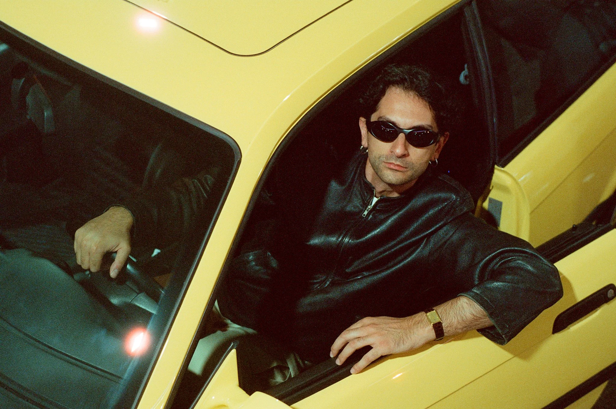 A man with dark, curly hair and sunglasses sits in a yellow vehicle, leaning out of a window and looking at the camera.