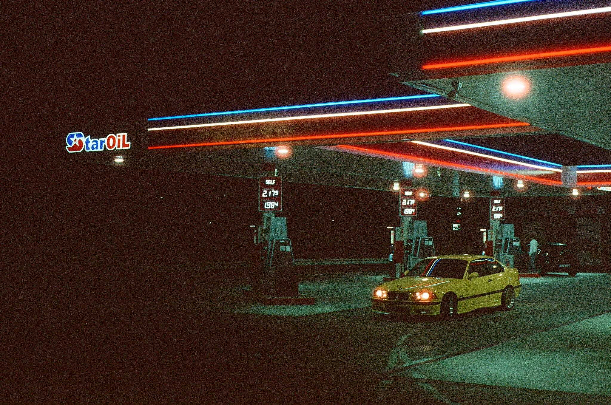 Night scene of a Star Oil gas station illuminated with neon lights in red, white, and blue. A yellow car is parked at the pump, and digital price signs are visible.