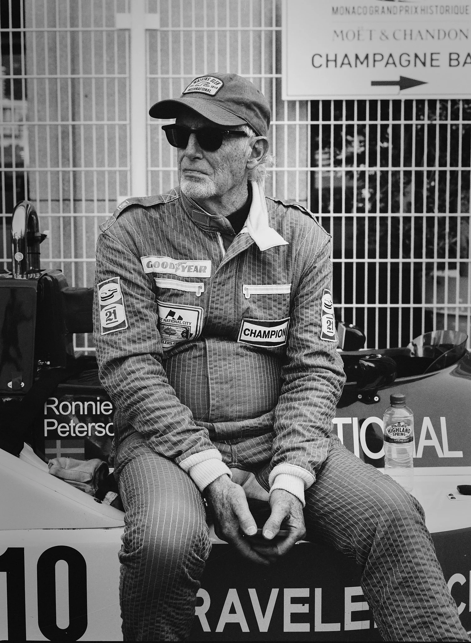 A man wearing sunglasses, a cap, and a patterned racing suit sitting outdoors near a metal fence, with a sign pointing to a champagne bar in the background, and a water bottle on the table nearby.