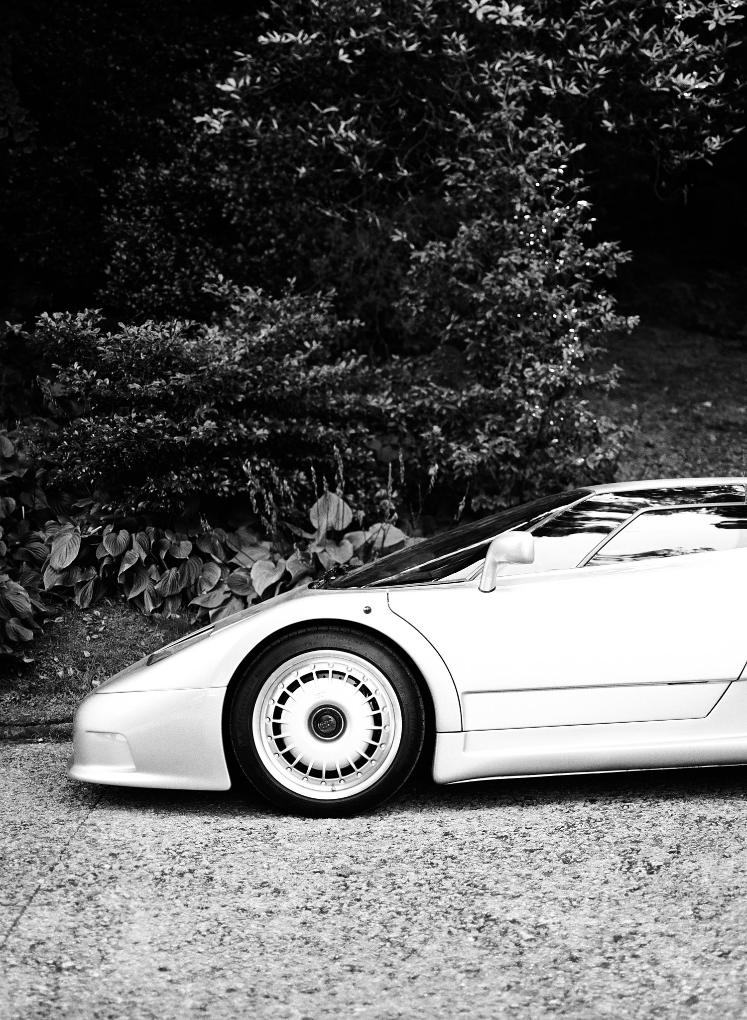 A partial view of a white sports car parked on a textured street, with a dark leafy background and the car's front wheel and part of the front fender visible.
