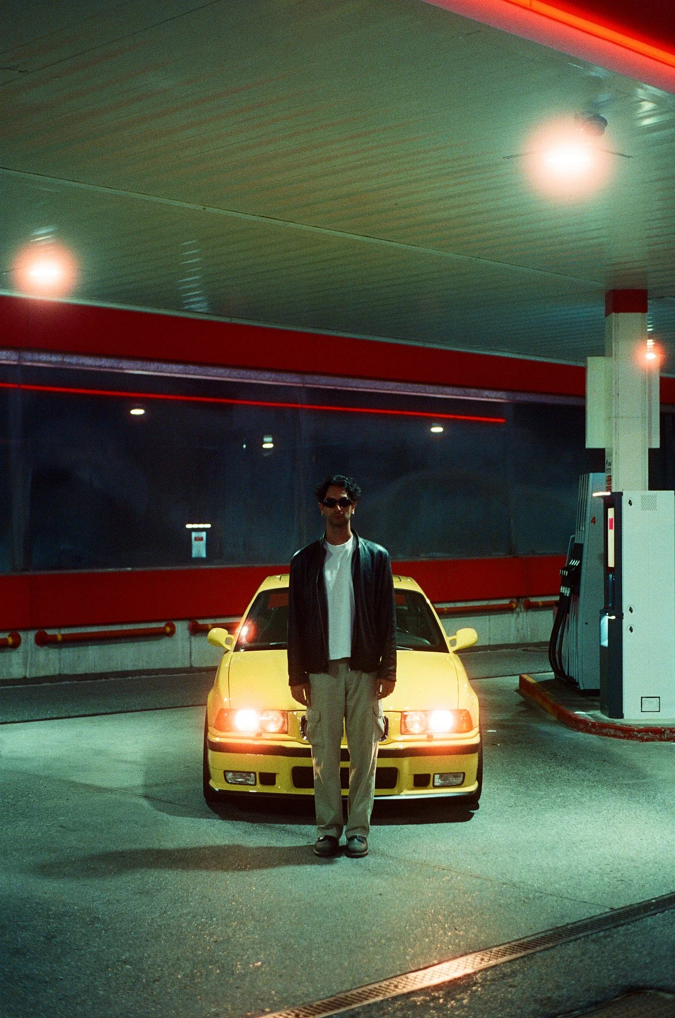 A man standing in front of a yellow sports car at a gas station at night, illuminated by overhead lights.