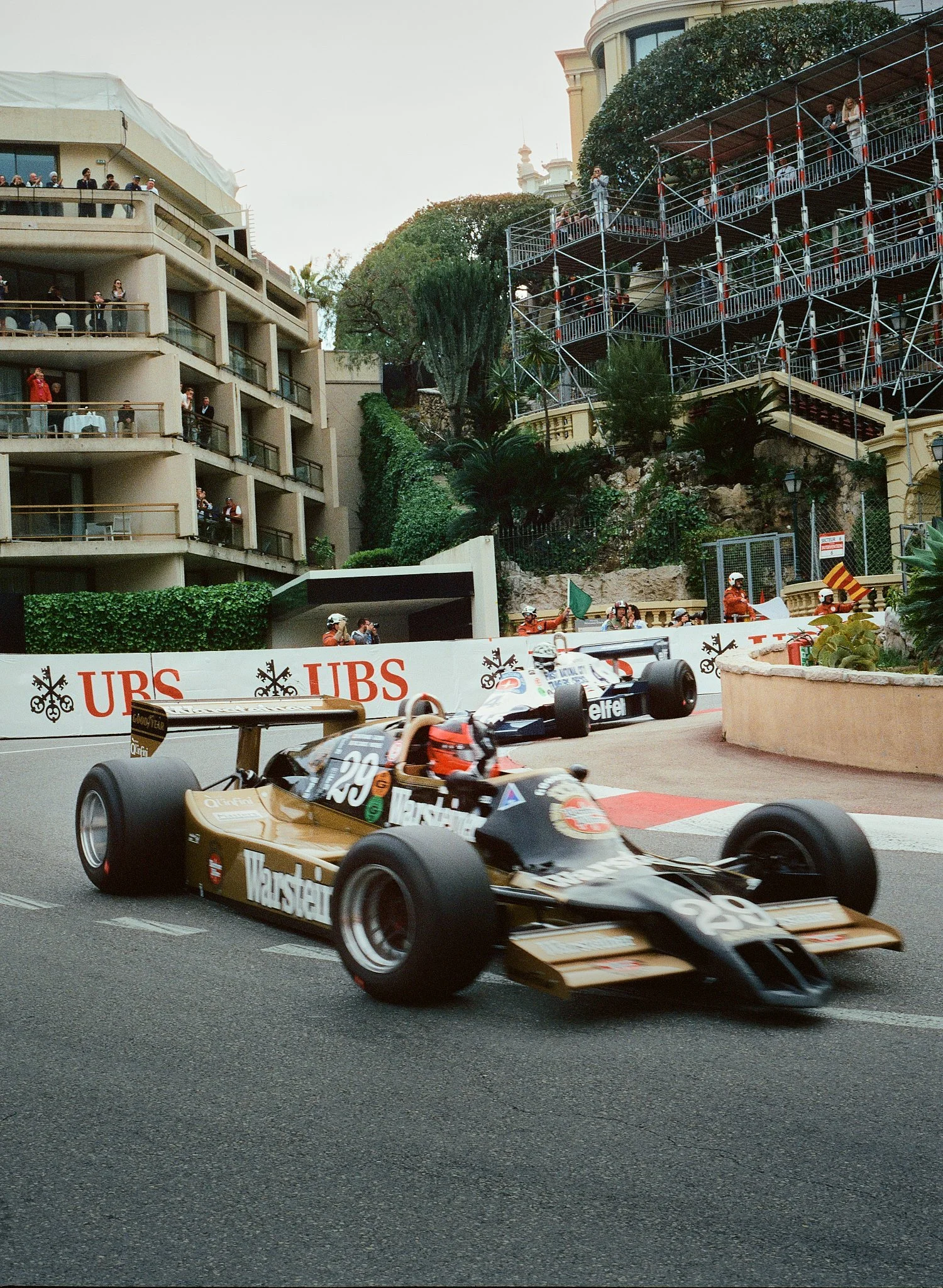 Race cars on a track with spectators watching from balconies and scaffolding, trees, and buildings in the background.