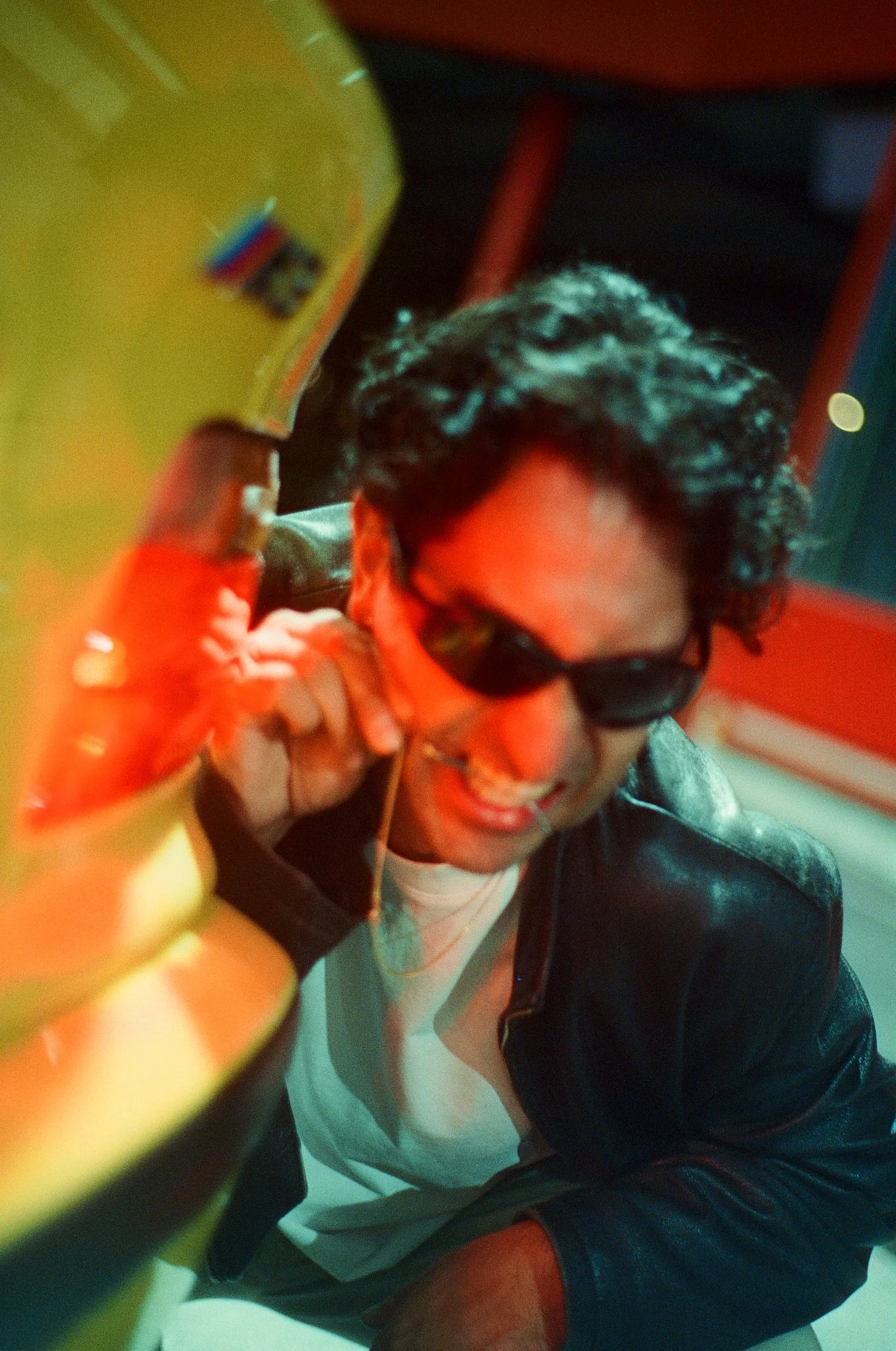 A man with curly hair and glasses smiling, posing with a peace sign in front of a yellow object, possibly a toy or a container, and wearing sunglasses and a black leather jacket.