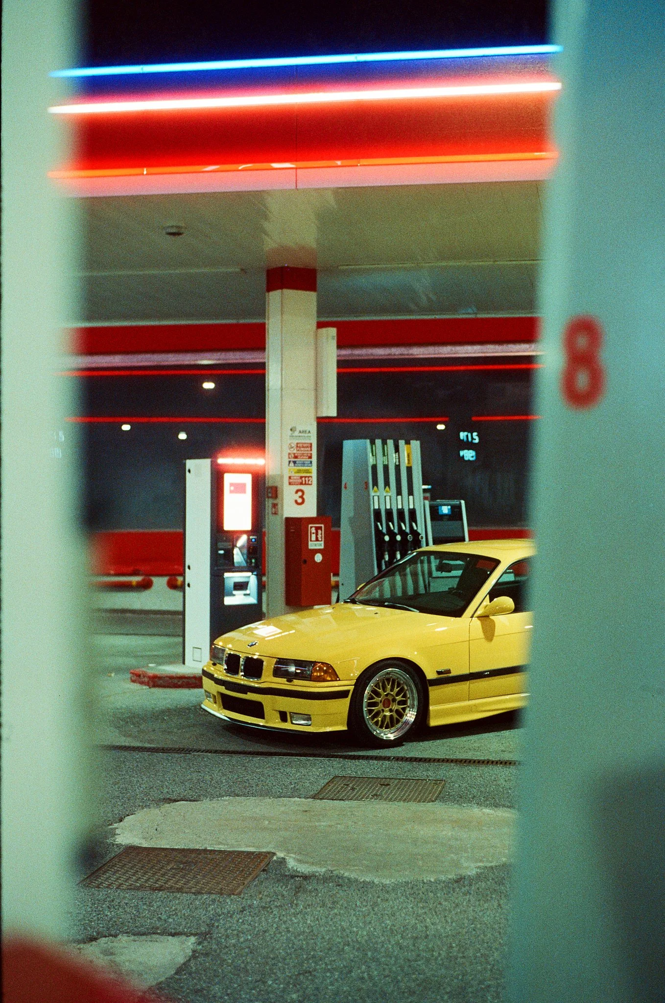 A yellow BMW car parked at a gas station entrance, viewed through a window with greenish partial borders.