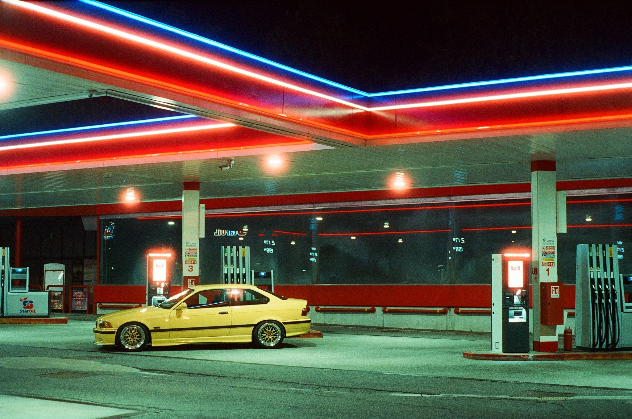 A yellow sports car parked at a gas station with neon red and blue lights illuminating the ceiling.