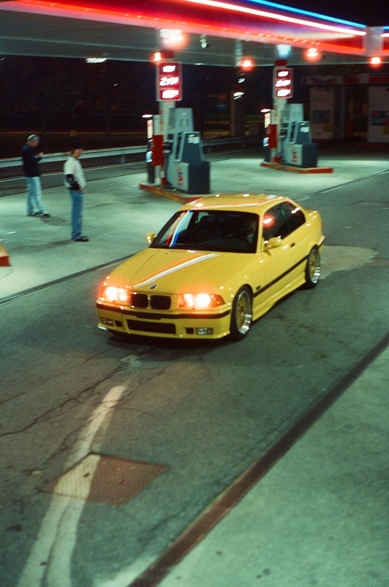 A yellow sports car parked at a gas station at night, with two people standing nearby and illuminated gas pumps under a brightly lit canopy.