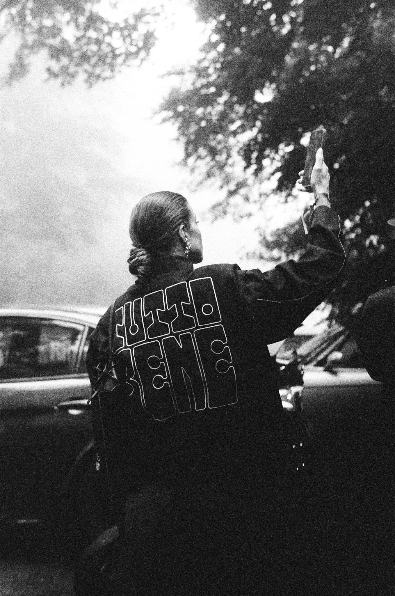 A woman with her hair tied back, wearing earrings and a jacket, holding a phone in her right hand as she takes a selfie outdoors, with trees and cars in the background, in black and white.