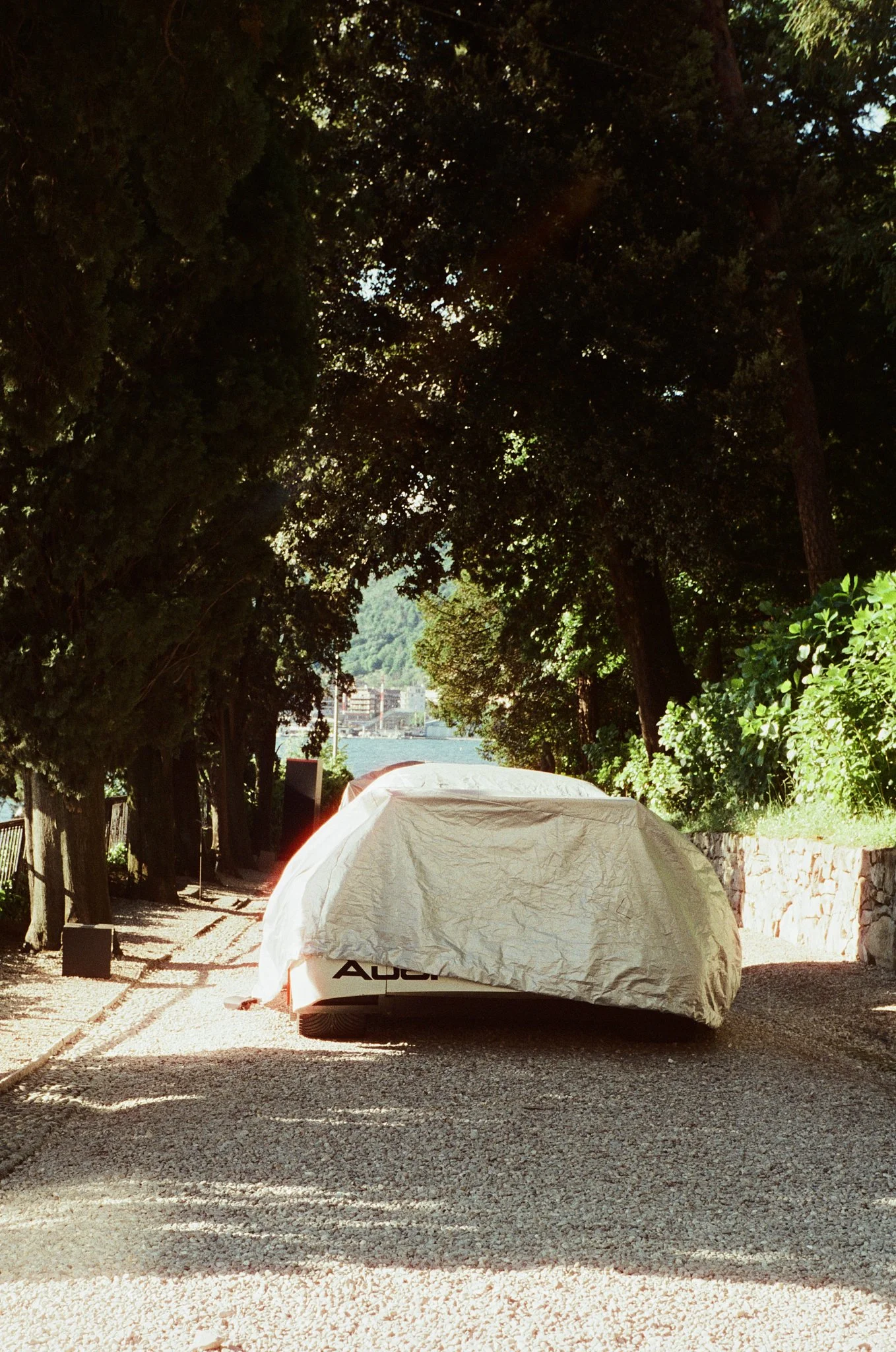 White covered car parked on gravel path with trees on either side and a view of water and buildings in the distance.