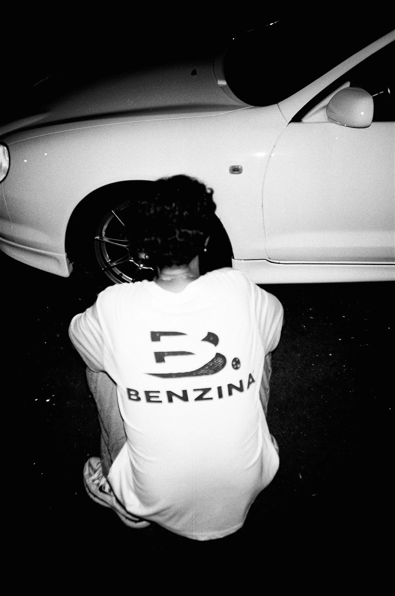 A person with curly hair wearing a white Benzina T-shirt, squatting in front of a white car at night, with their back facing the camera.