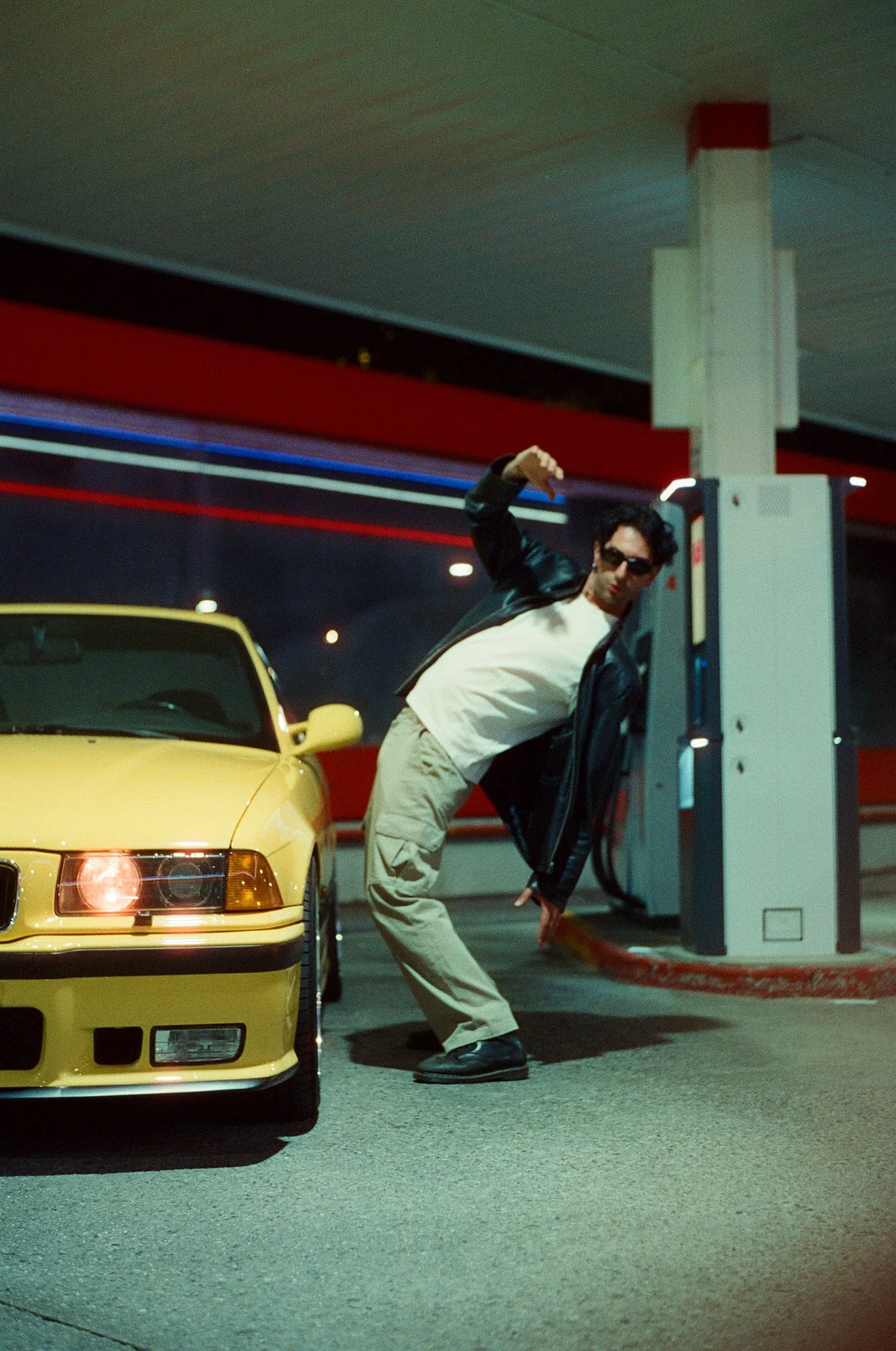 A man in a black jacket, sunglasses, and beige pants is at a gas pump, bending down and reaching with one hand while the other hand is raised above his head in a pose. A yellow sports car is parked nearby, and there is a gas station canopy with red a