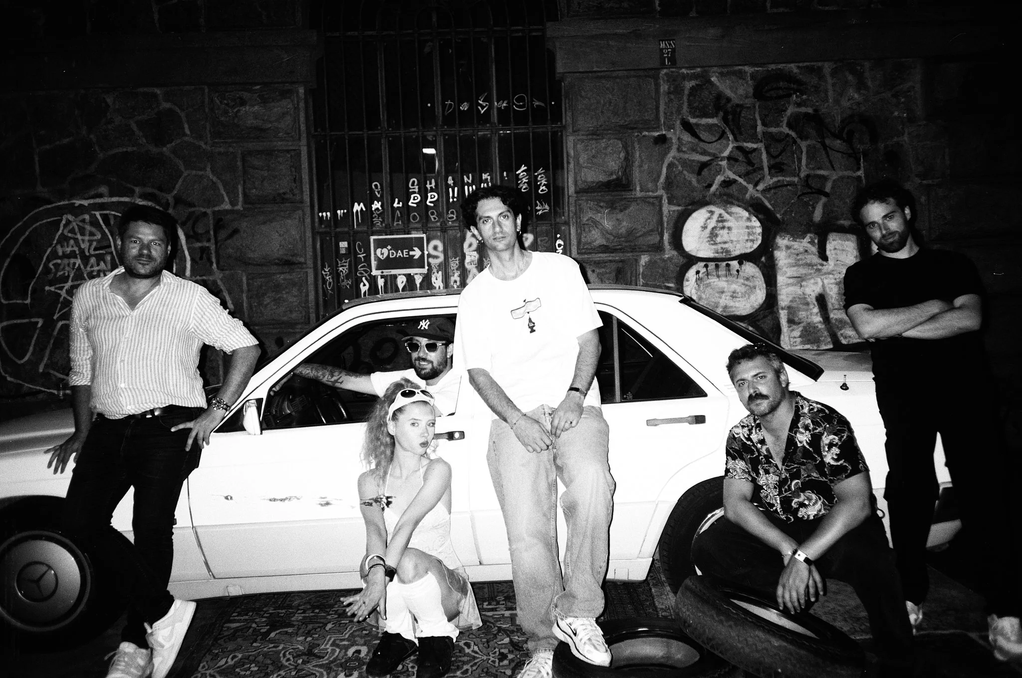 A group of six young adults, including four men and two women, posing in front of a white car on a street at night with a graffiti-covered wall in the background.