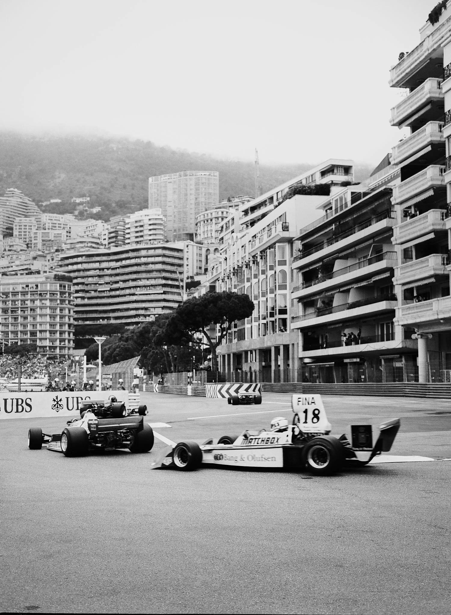 Black and white photo of a Formula 1 race track with racing cars moving at high speed and buildings with balconies and trees in the background.