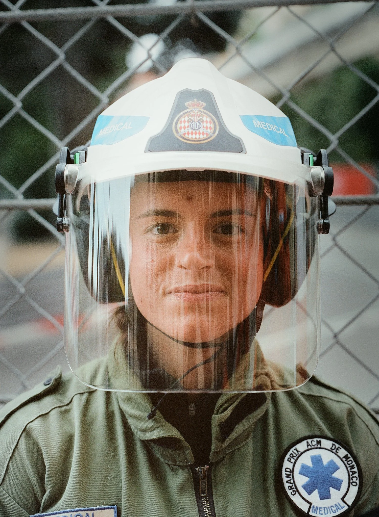 A person wearing a uniform with a medical patch and badge, and a safety helmet with a face shield and medical insignia, standing behind a chain-link fence.