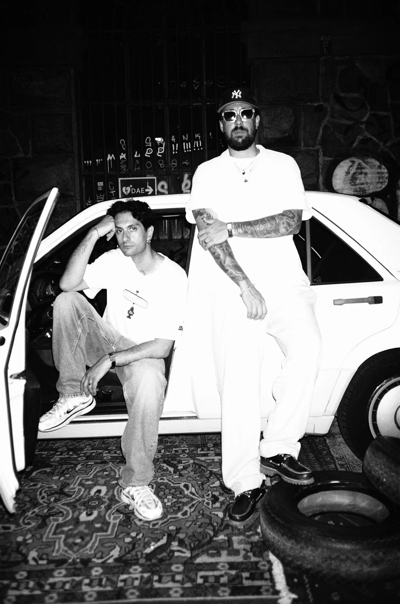 Black and white photo of two men posing with a vintage car in an indoor setting. One man is sitting with his arm resting on his head, wearing a white t-shirt, jeans, and sneakers. The other man is standing, wearing a white t-shirt, white pants, sungl