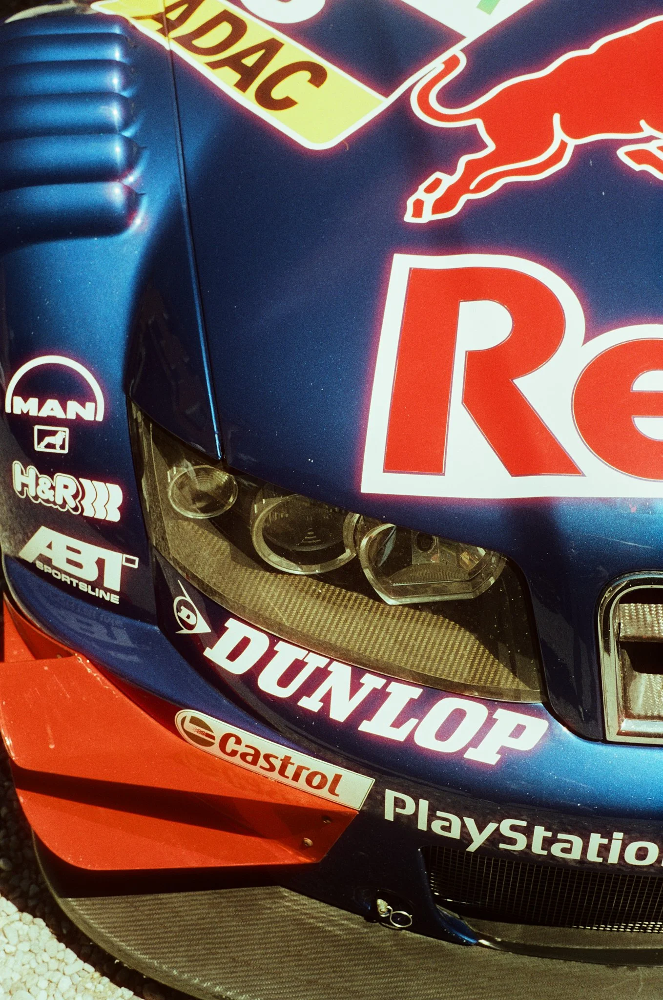Close-up of a race car's front, featuring various sponsor decals including Castrol, PlayStation, and others. The car has a blue and red color scheme.