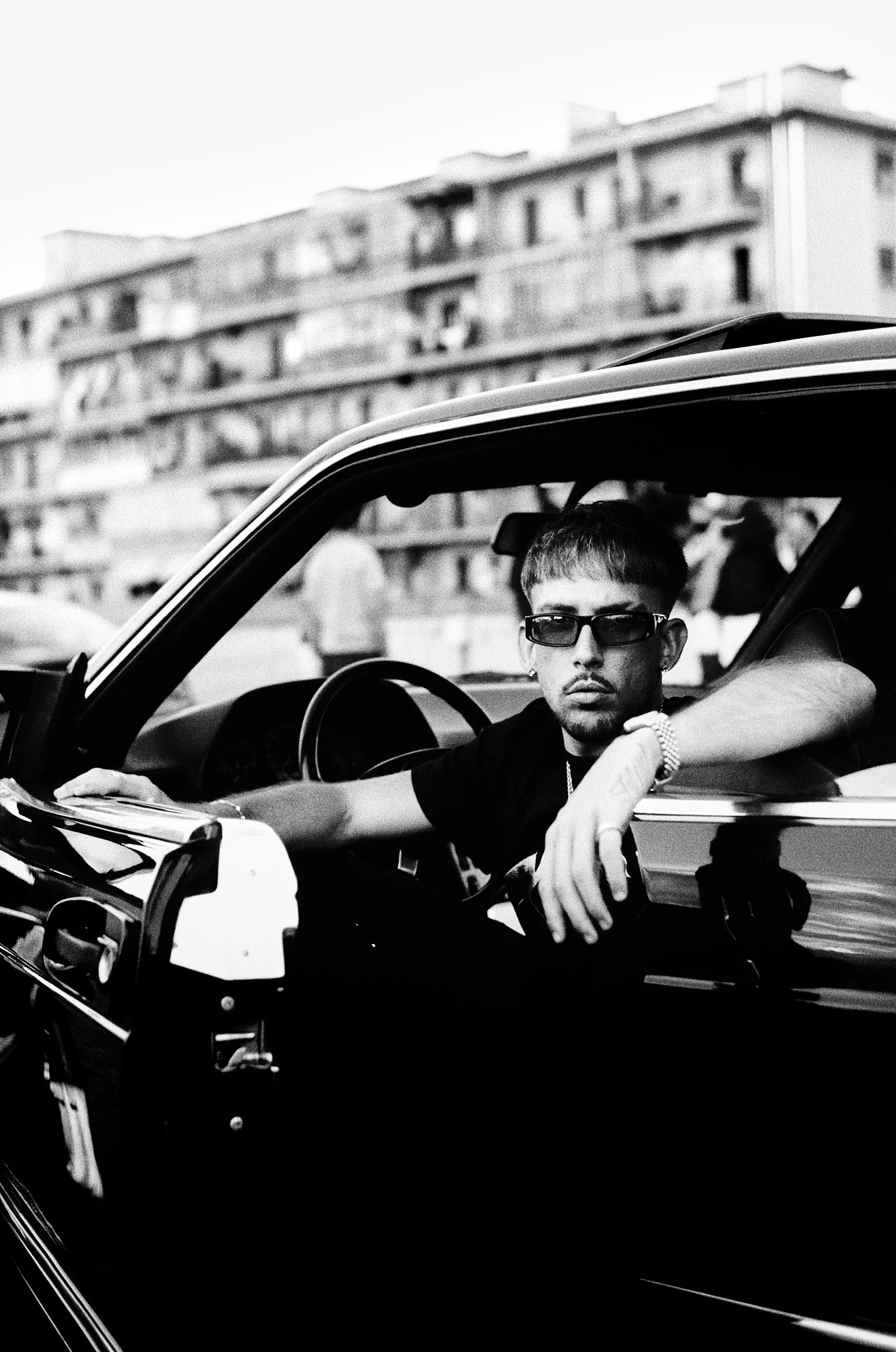 A man with sunglasses sitting in a car with his arm resting on the window. A building is blurred in the background.