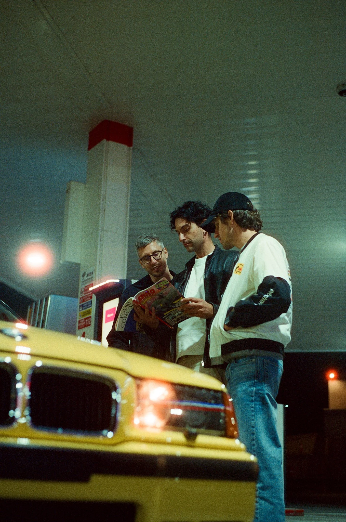 Four men standing near a yellow taxi at a parking garage, looking at a map or magazine.