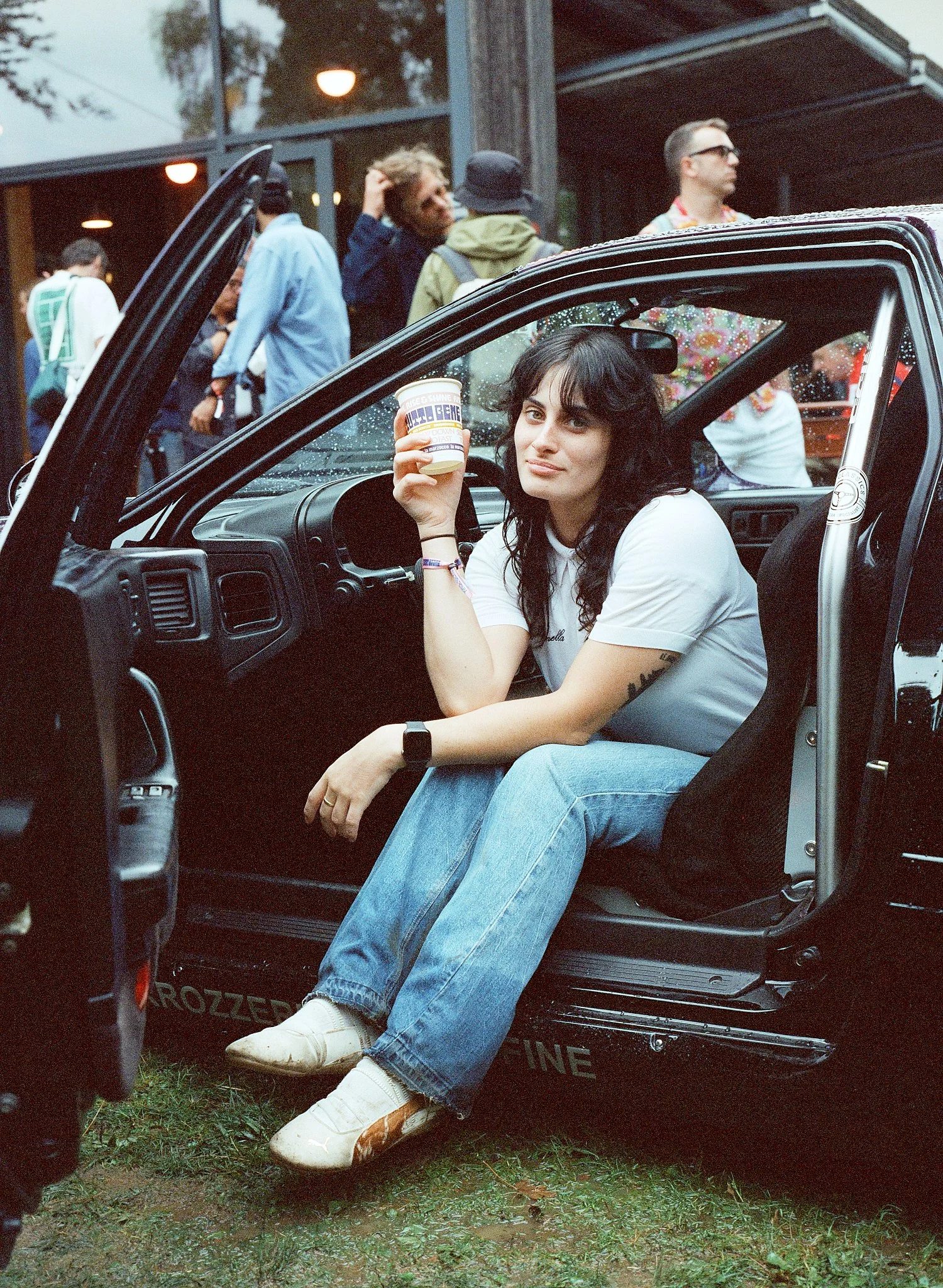 Woman sitting in the driver's seat of a black vehicle, holding a container of ice cream, with a slight smile, wearing a white t-shirt and blue jeans. Other people are standing outside the vehicle, talking in a casual outdoor setting, with trees and a