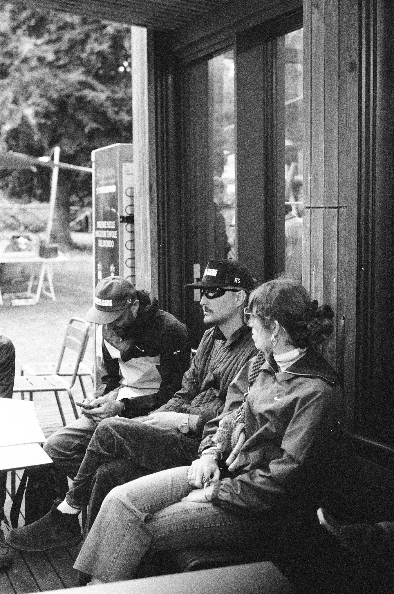 Four people sitting at a row of chairs outside a building, with a vending machine and outdoor scenery in the background. One person is looking at a phone, while the others are facing forward.