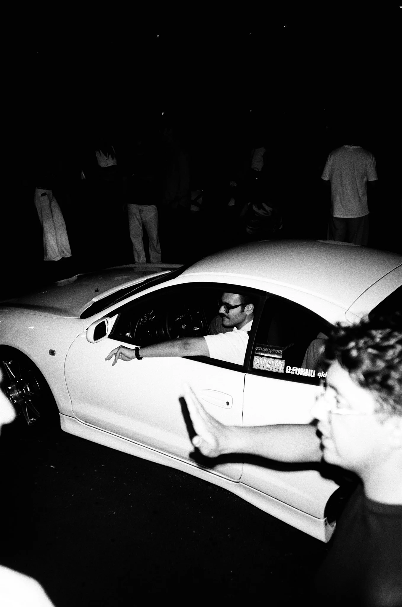 A man wearing glasses sits in a white sports car, smiling and extending his arm with his hand pointing. In the background, there are several people standing, some with crossed arms, in a dark outdoor setting at night.