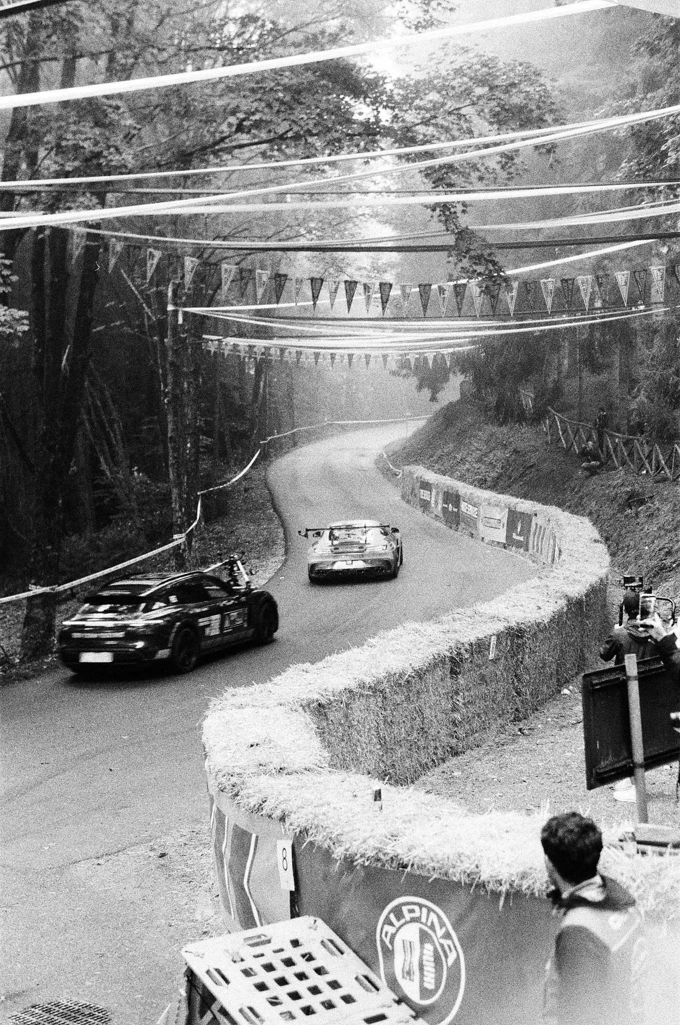 A street scene with a race car on a winding road surrounded by trees. There is a person in the foreground wearing a jacket with a logo, looking towards the race car, and a barrier with a basket and a sign on the sidewalk.
