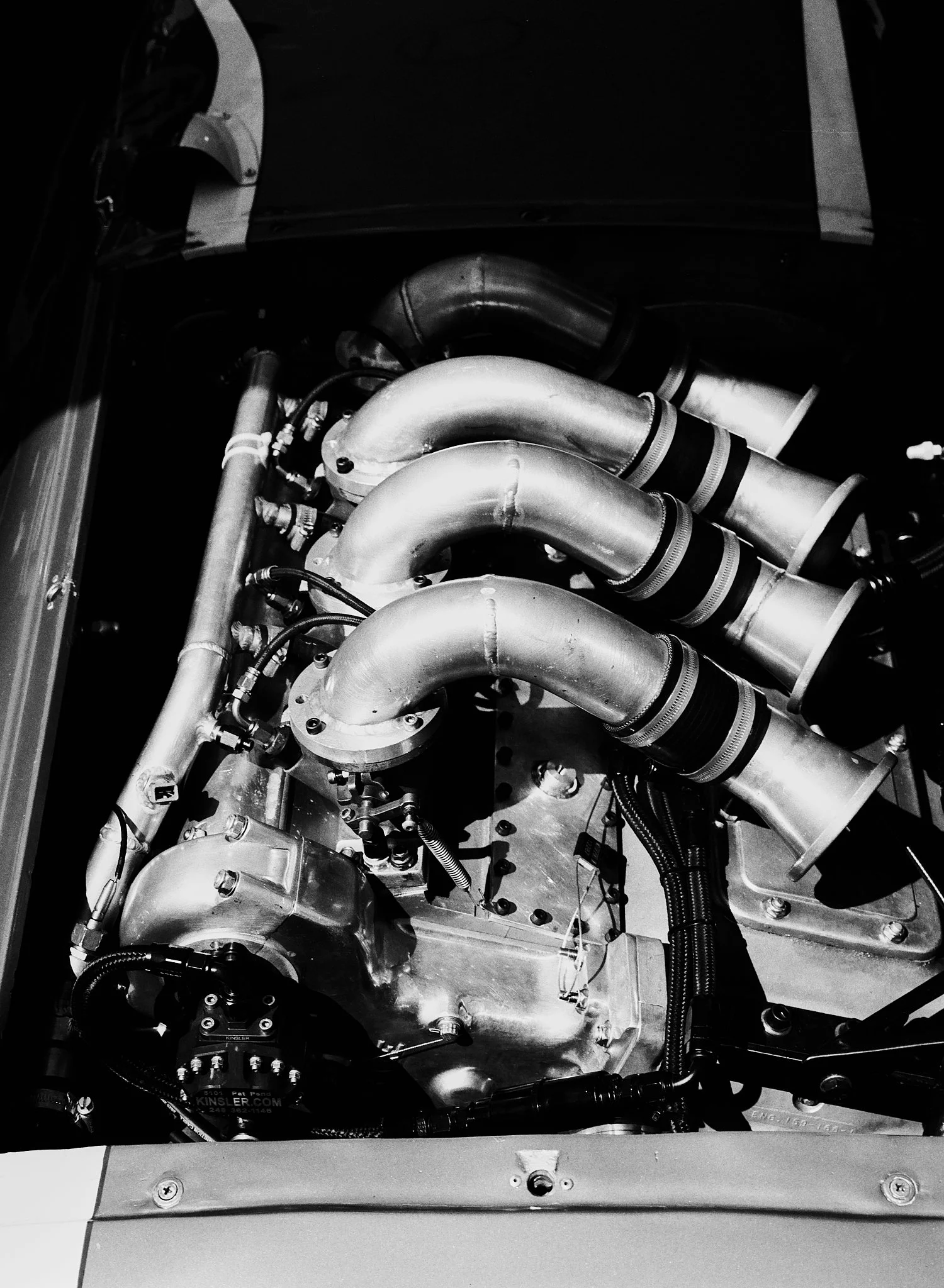 Black and white photo of a car engine with metal pipes, hoses, and mechanical components.