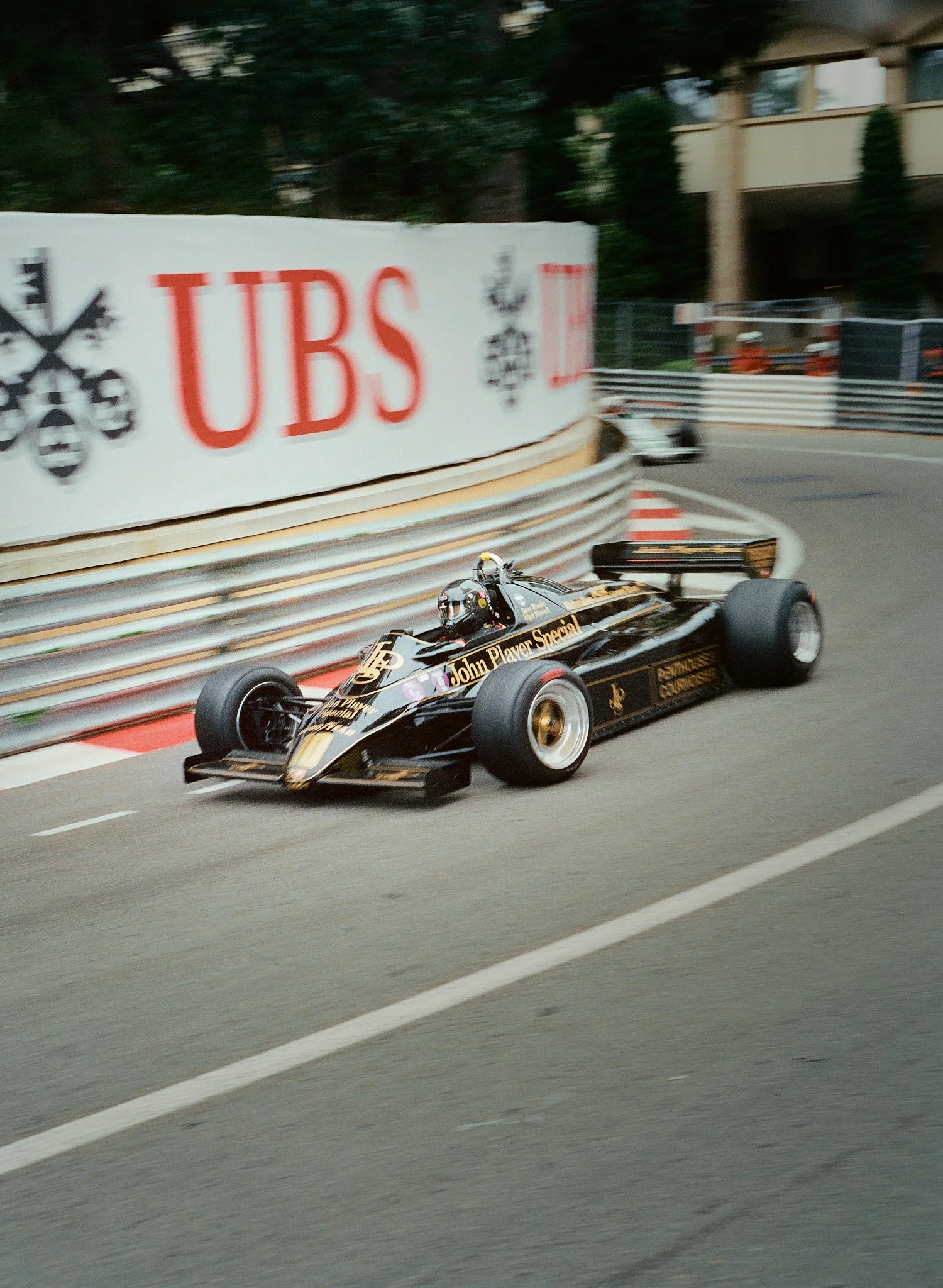 A black Formula 1 race car with gold accents, sponsored by John Player Special, navigating a curve on a race track with safety barriers and a large advertisement for UBS in the background.