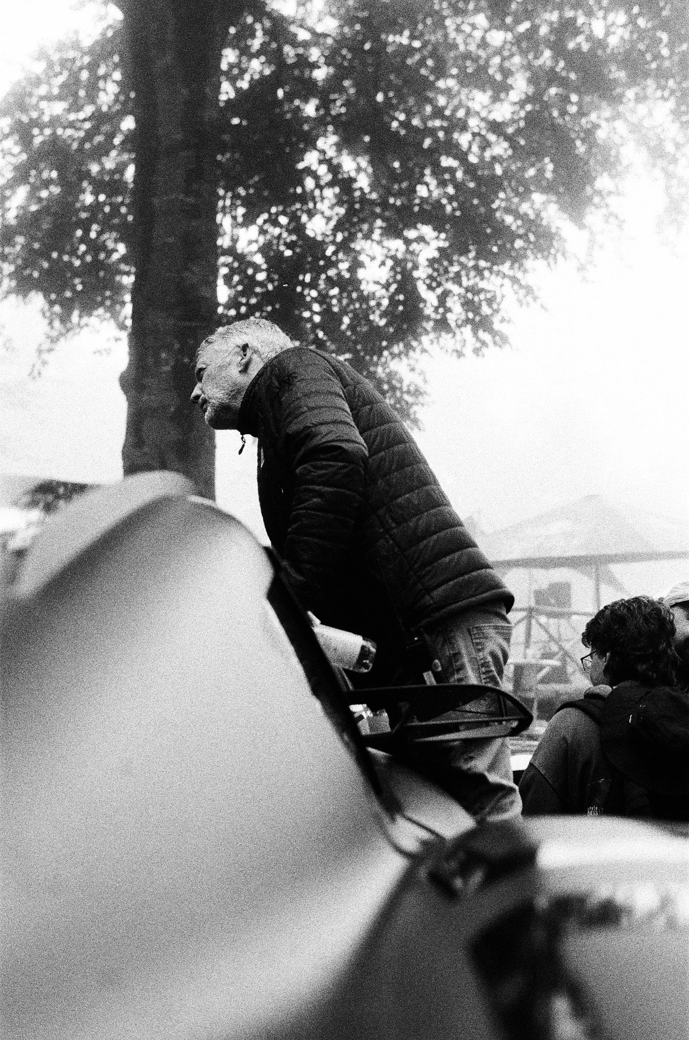 Black and white photo of several people outdoors with large trees in the background. One man wearing a quilted jacket and glasses appears to be looking to the side, surrounded by others, at what seems to be a gathering or event.