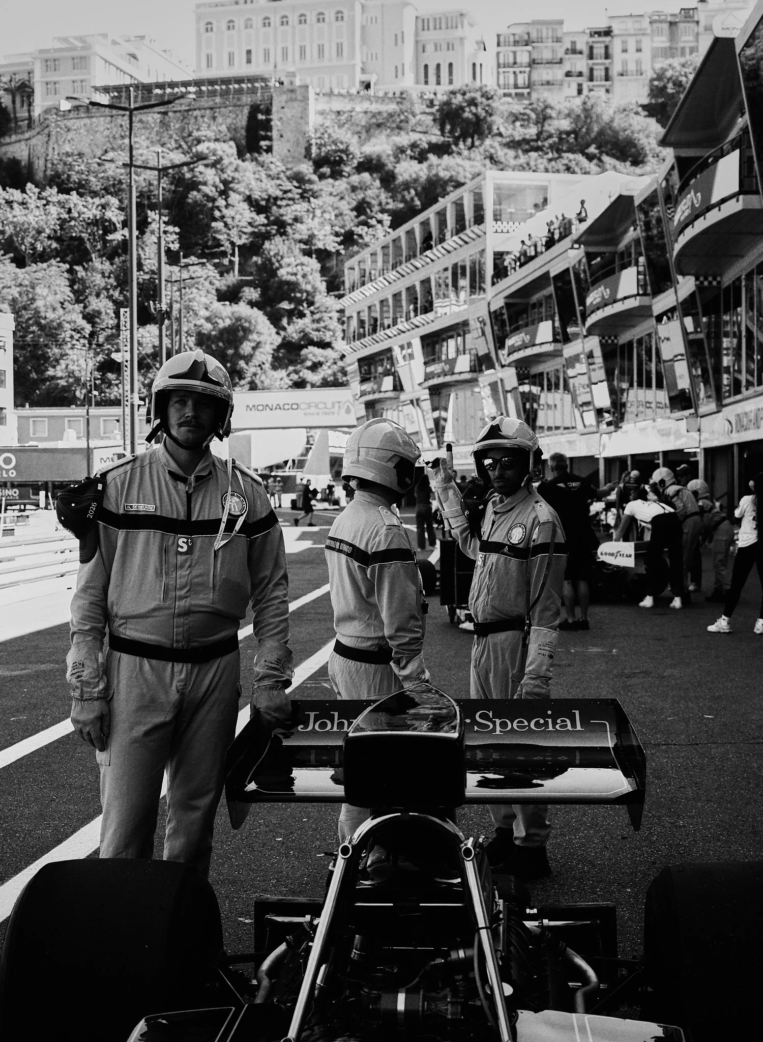 Four race officials in racing uniforms and helmets standing near a Formula 1 car on a city street circuit, with tall buildings and audience stands in the background.