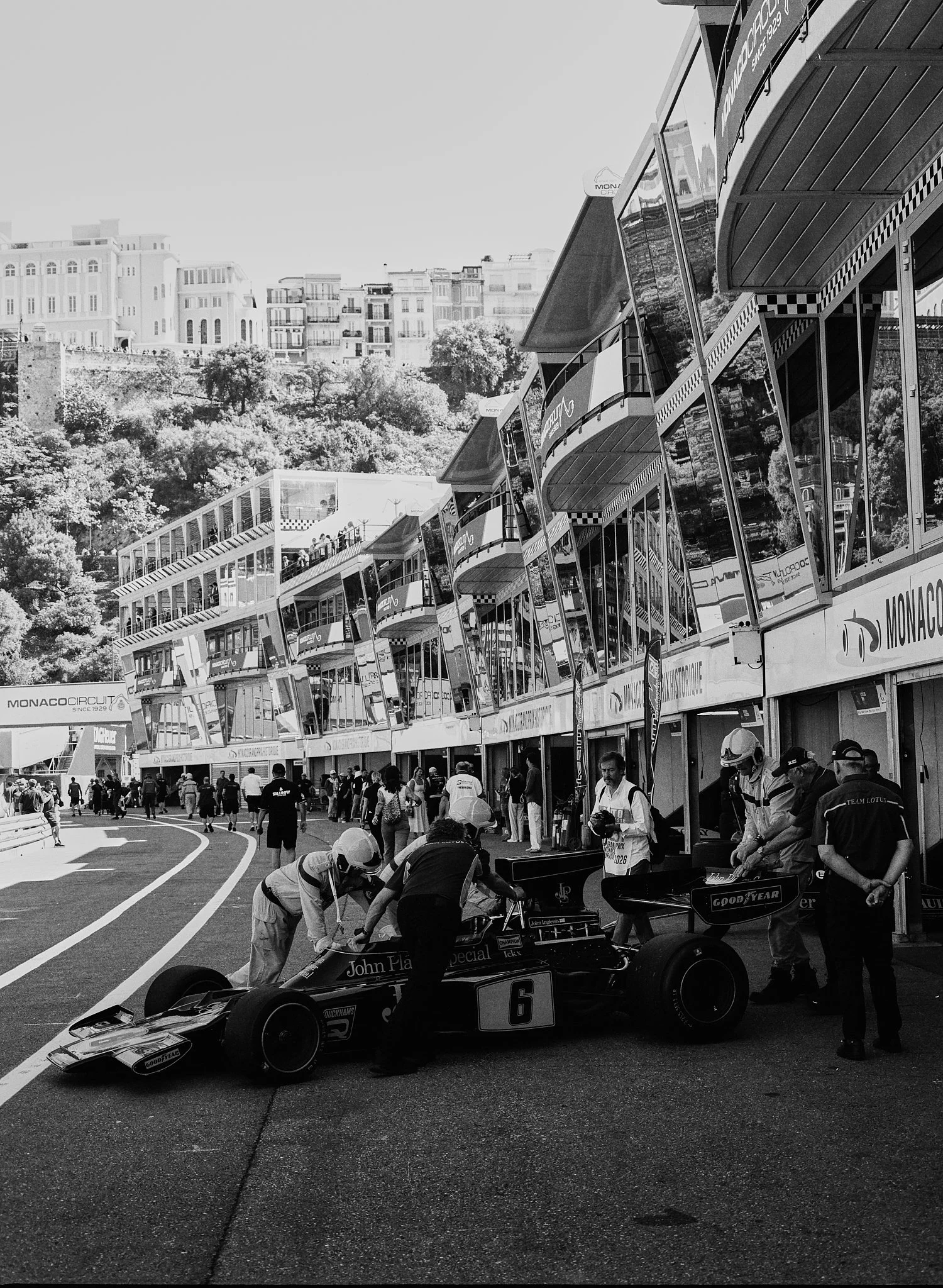 A black and white photo of a racing pit area at the Monaco Grand Prix. Several race cars are being serviced by mechanics, with one car prominently in the foreground bearing the number 6. People are walking and working near the cars, and there are mul