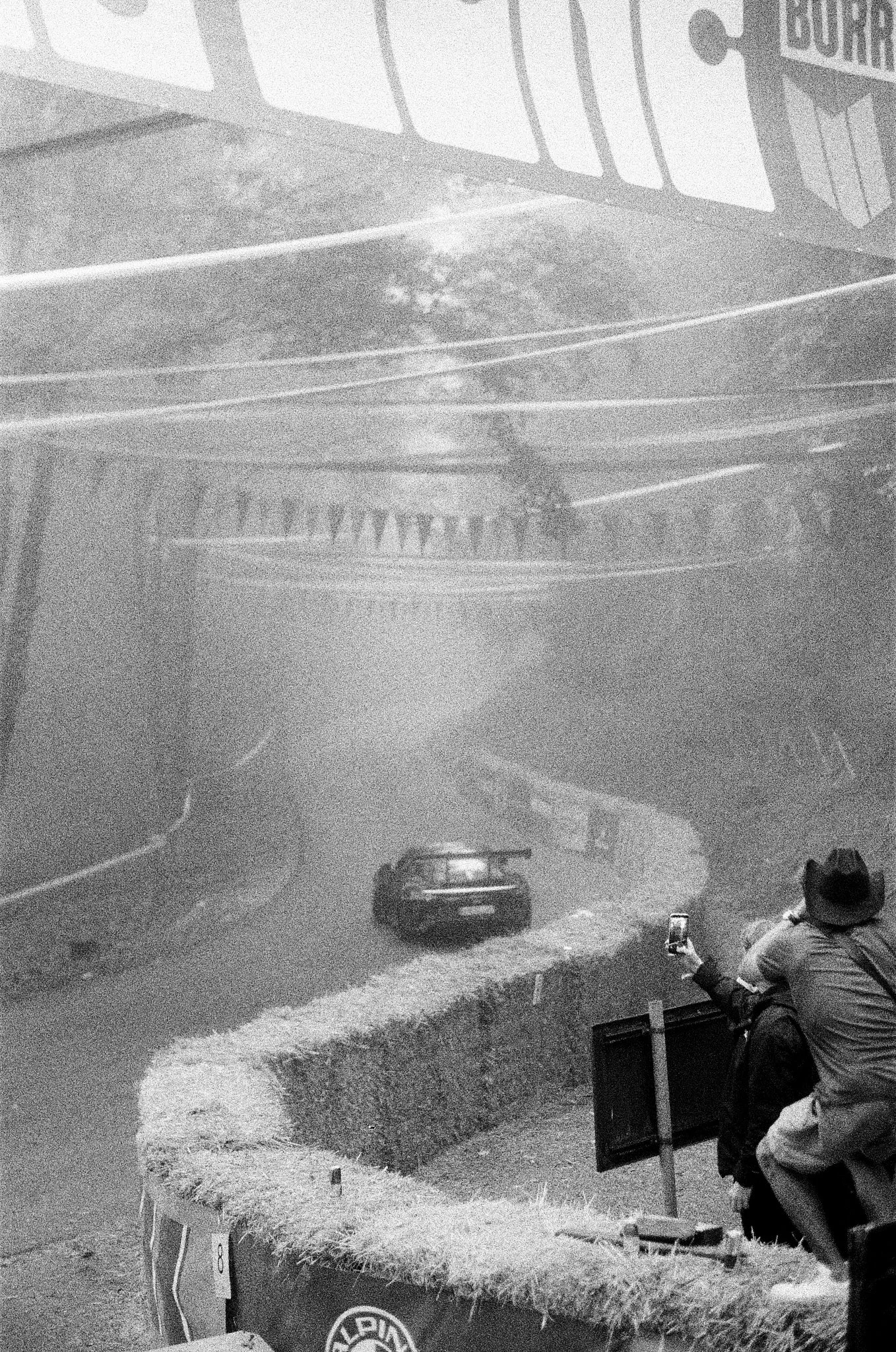 Nighttime scene of a highway with a black car driving and a person sitting on a roadside fence taking a photo of the car. There are road signs along the road and a barrier with grass and plants on the roadside.