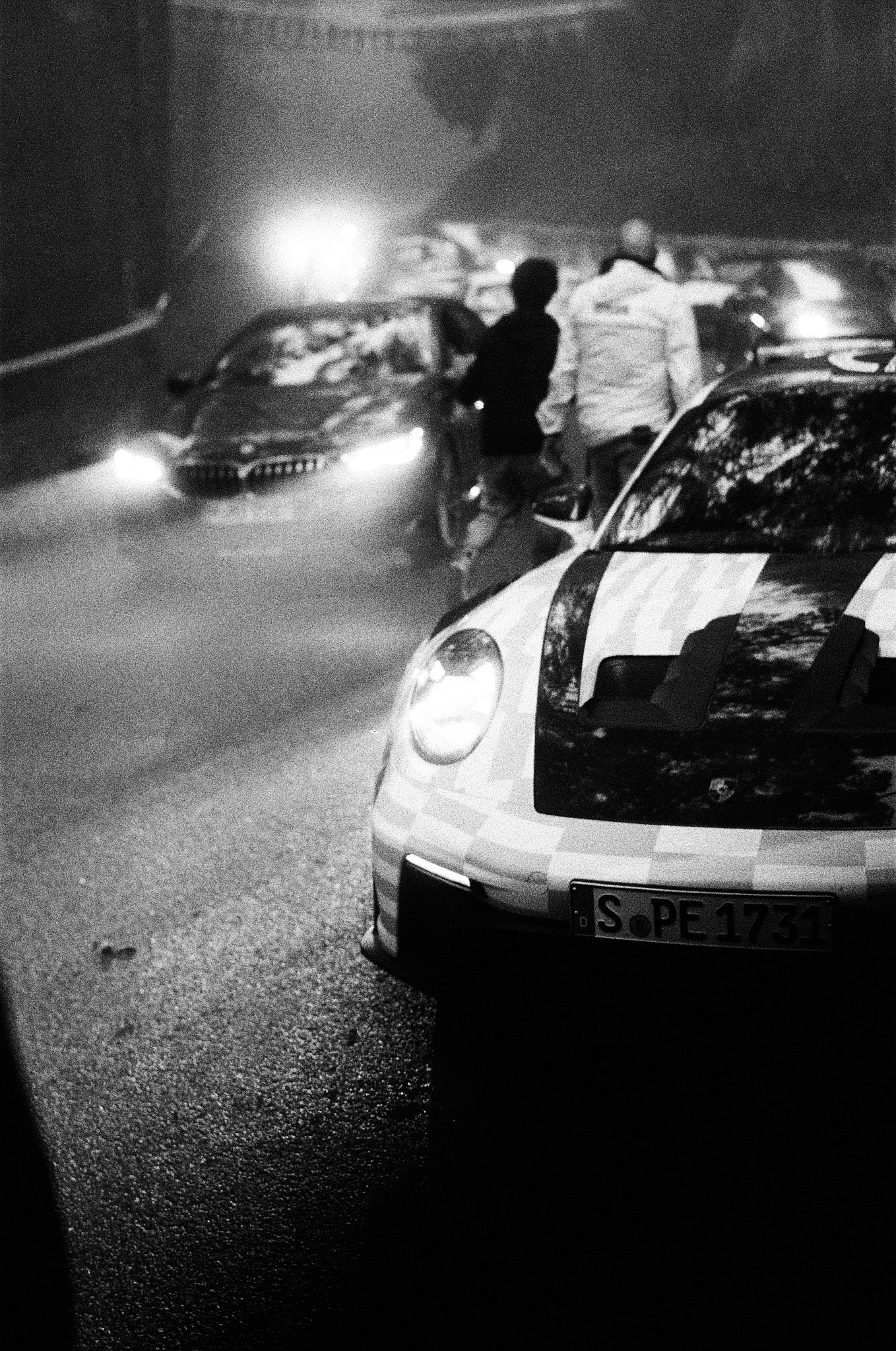 Nighttime scene showing police cars and two individuals standing by a disabled car on the roadside, with blurred headlights and wet pavement.