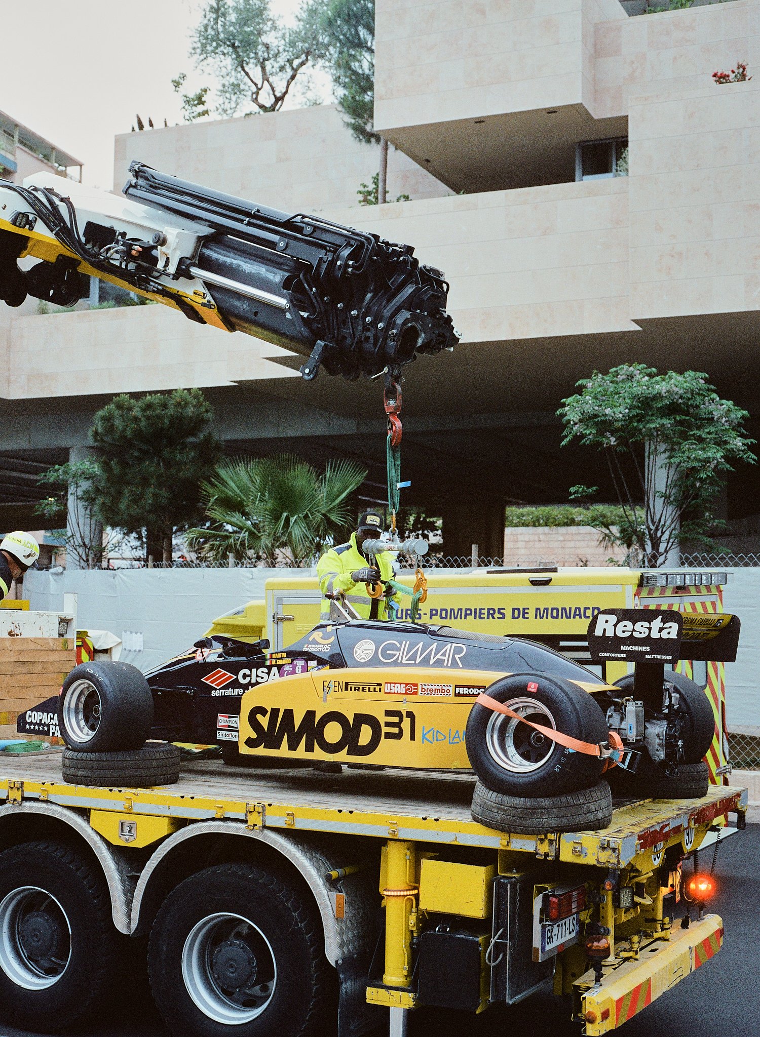A yellow tow truck carrying a Formula 1 race car with sponsorship decals, surrounding equipment, and workers in safety gear at an outdoor location.