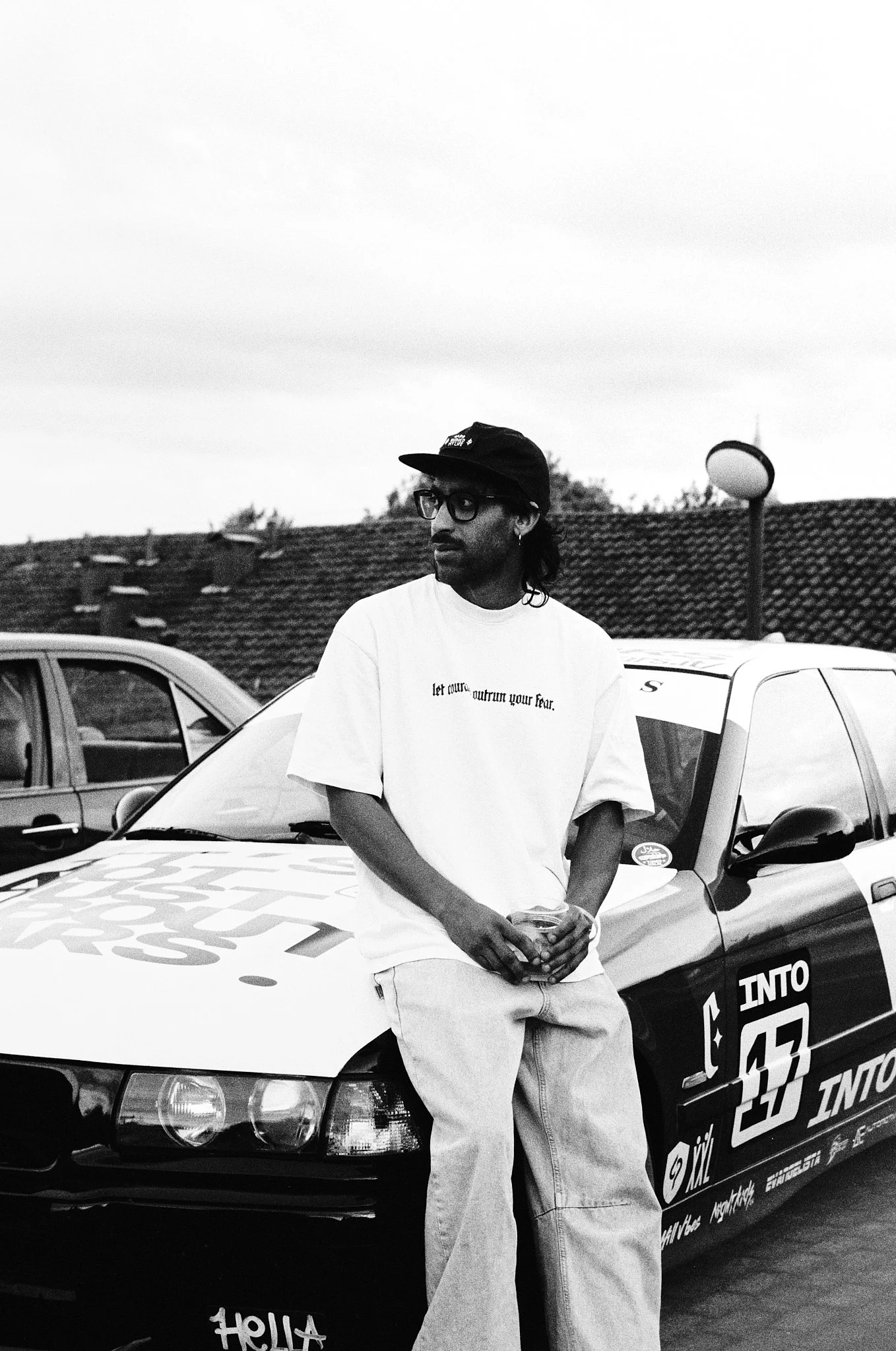 A man with glasses and a black cap stands in front of a race car. He is holding a drink and wearing a white t-shirt and baggy pants. The car has various sponsor logos and a prominent number 55 on the hood.