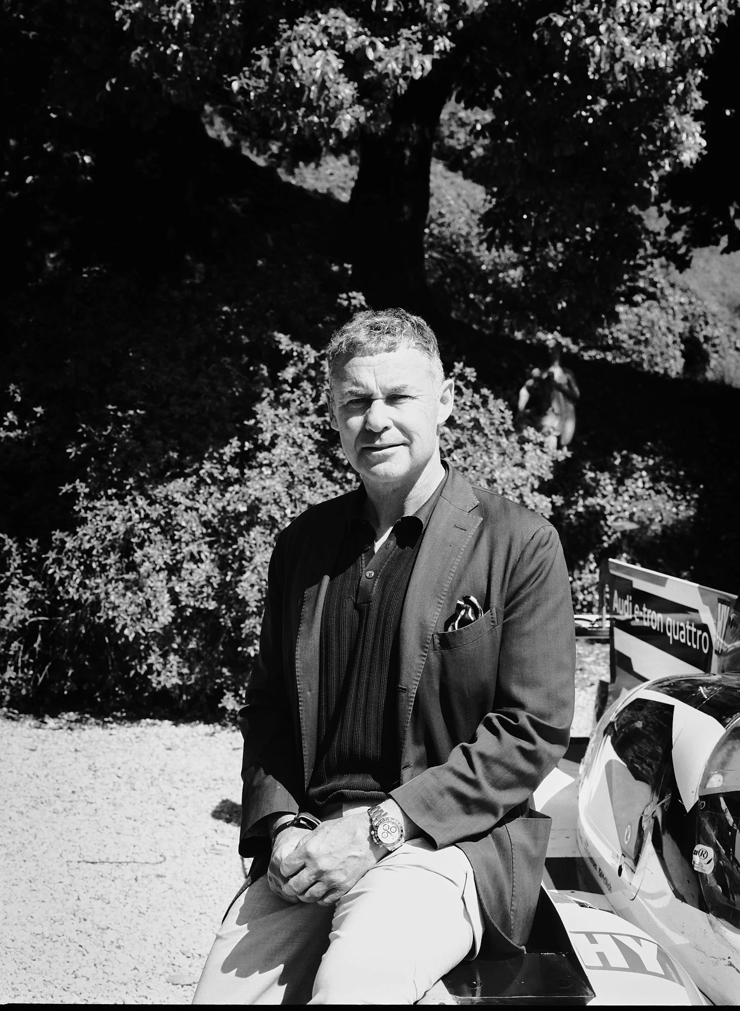 A man wearing a suit jacket and light-colored pants is sitting outdoors, with a tree and foliage in the background and a small race car nearby.