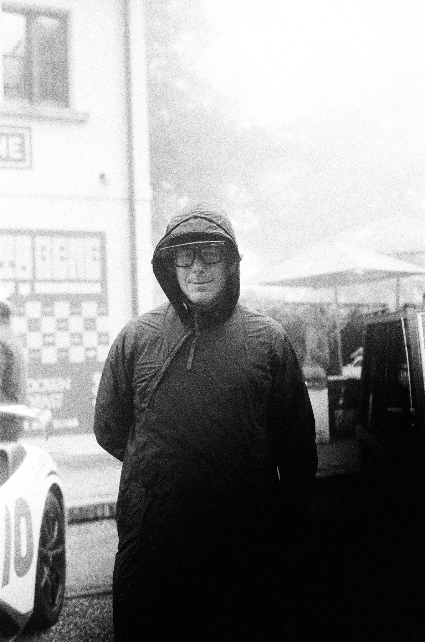 A man wearing glasses and a hooded jacket, standing outdoors in bad weather, with parked cars and a building in the background, and smoke or fog surrounding him.