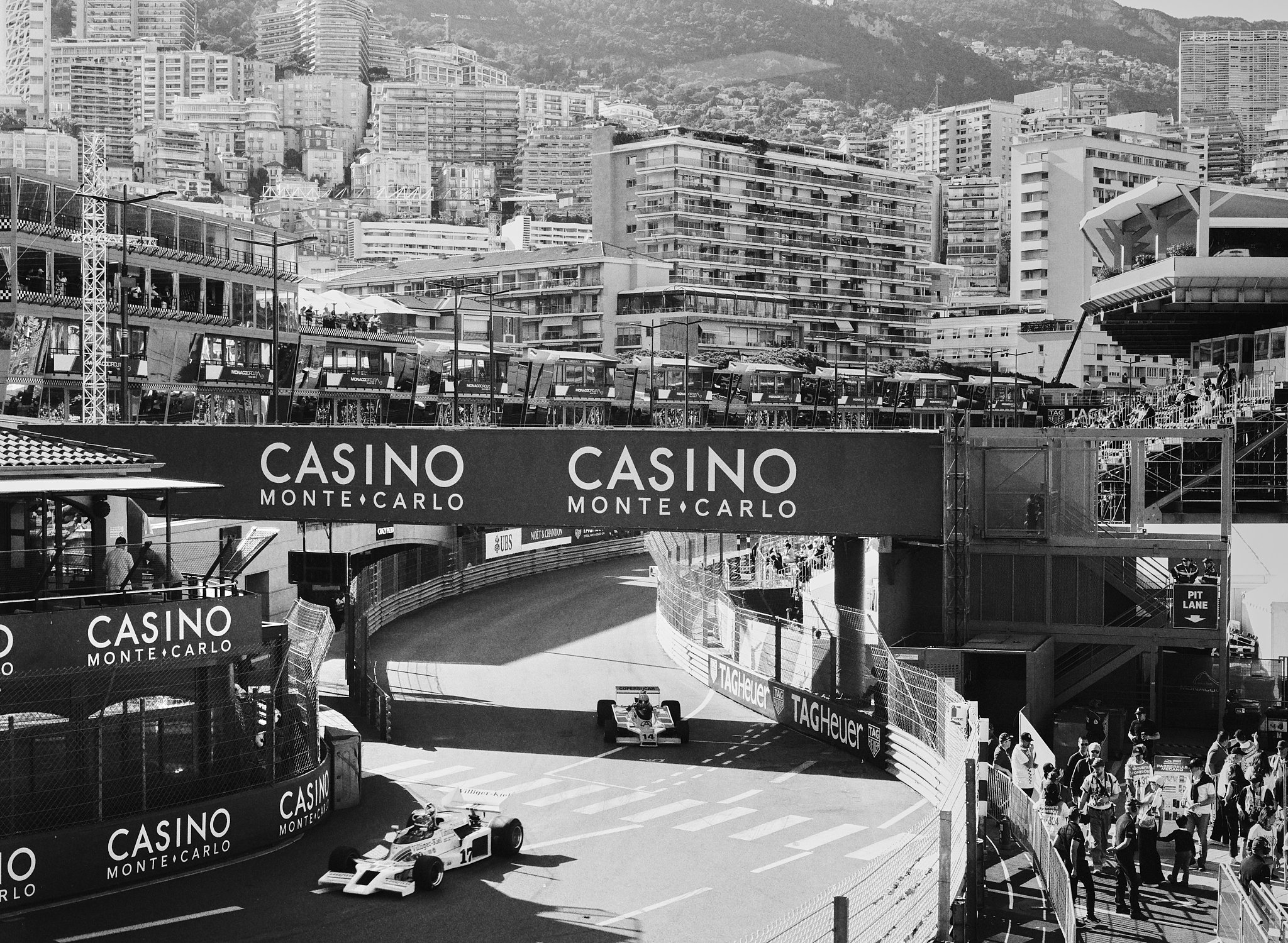 A black and white photo of the Monaco Grand Prix racing event with Formula 1 cars on the circuit, grandstands, and spectators. A large sign reads 'CASINO MONTE CARLO'.