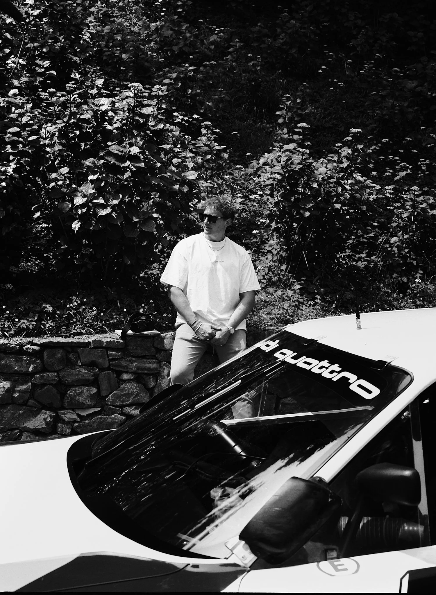 A man wearing sunglasses and a white t-shirt standing next to a police car with a roof-mounted antenna, in front of a backdrop of dense, leafy bushes.