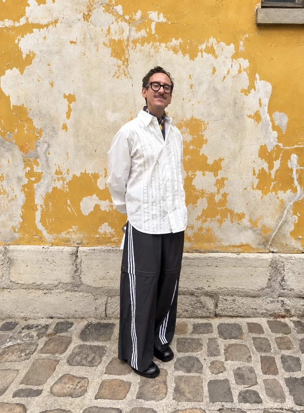 Un homme souriant portant des lunettes, une chemise blanche et un pantalon ample avec des bandes blanches, se tient devant un mur usé jaune et blanc, sur un sol pavé.