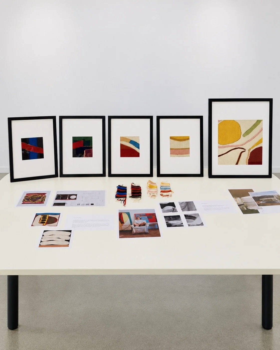 Exposition artistique avec cinq tableaux abstraits avec des formes colorées, placés en ligne sur un mur blanc, et divers échantillons de tissus, photos, et documents sur une table blanche au premier plan.