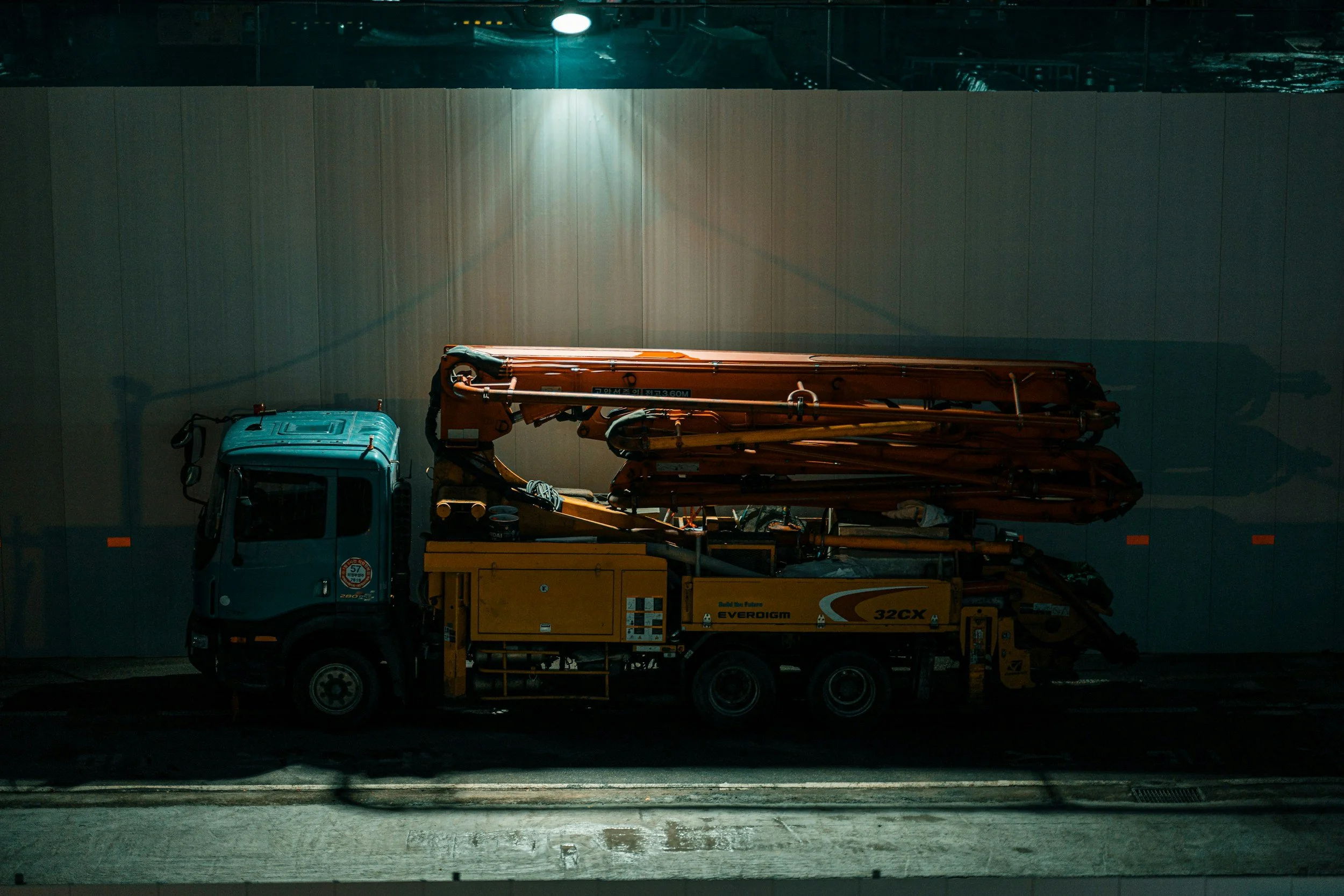 A truck parked in front of a large wall illuminated by nearby lights at night, carrying construction equipment including an orange boom lift and a yellow scissor lift.