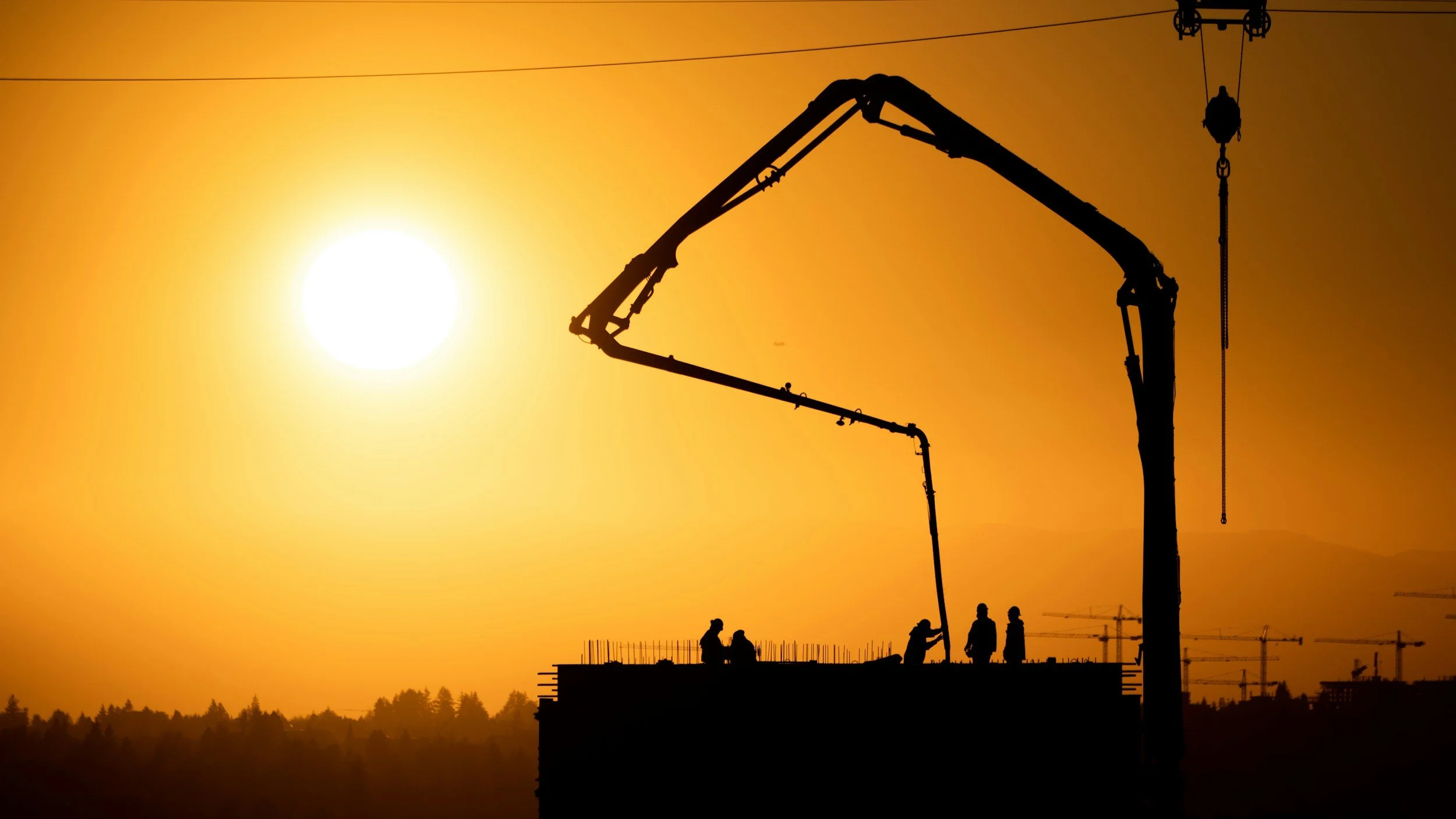 Construction workers standing on a building under construction against a bright orange sunset with silhouettes of cranes and construction equipment.