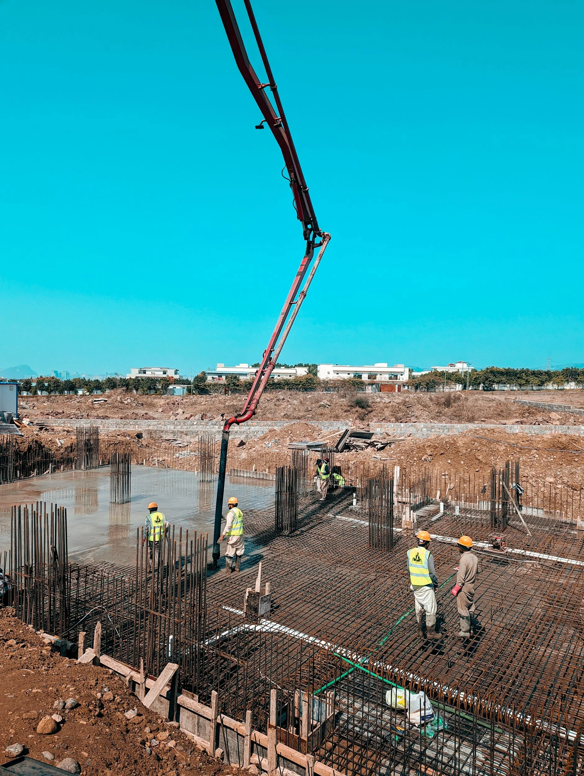 Construction workers in safety gear working on a building foundation with rebar and concrete, under a blue sky with industrial and residential buildings in the background.