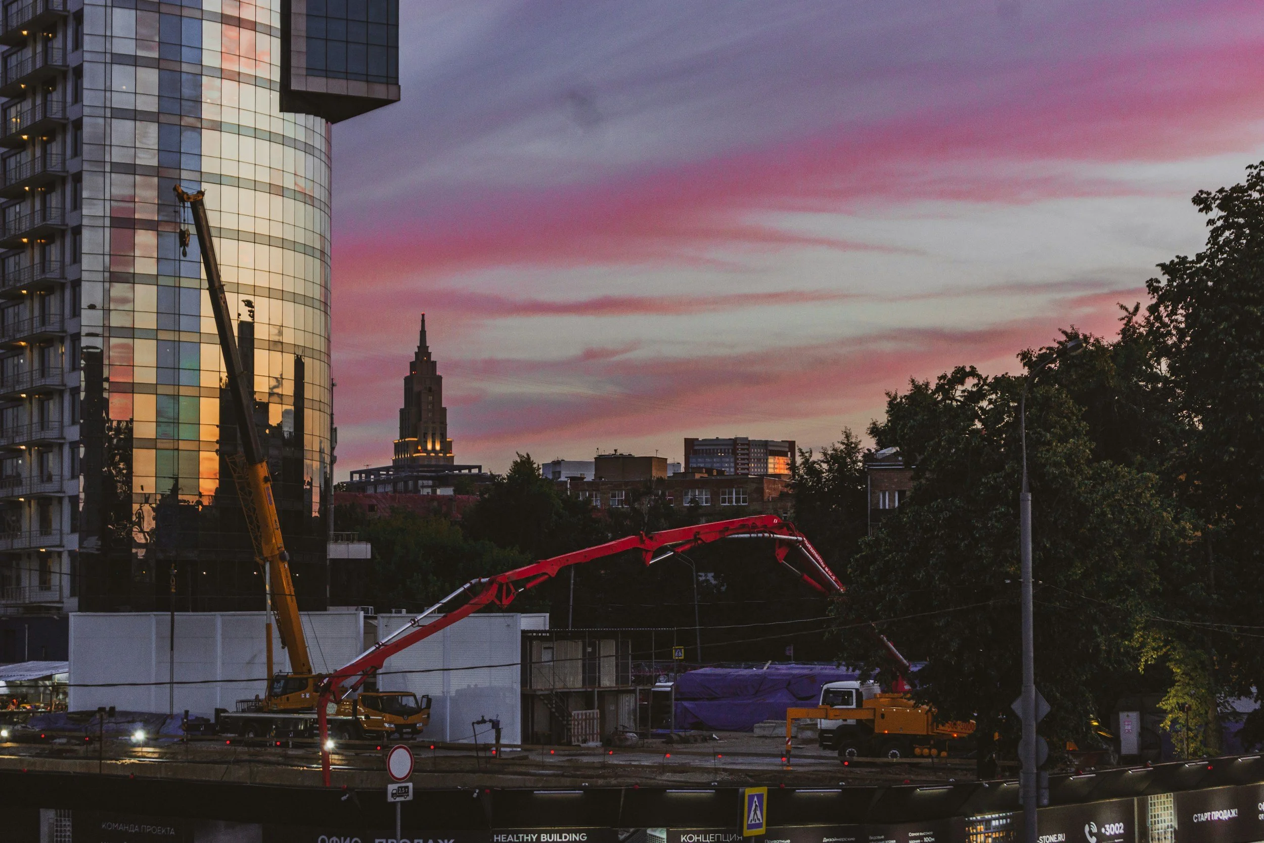Cityscape during sunset with modern glass buildings, construction crane, and a partly cloudy sky with pink and purple hues.