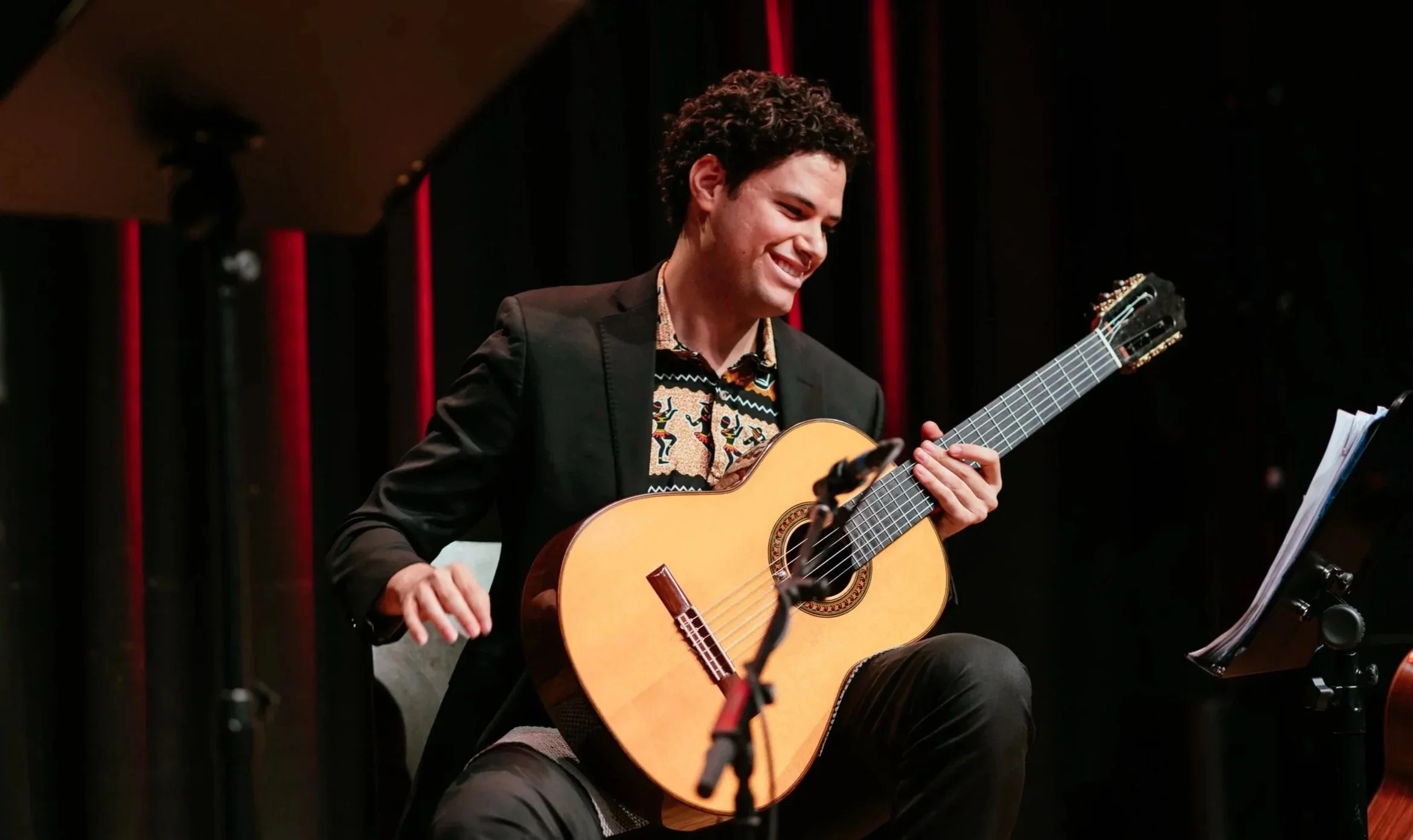 Junger Mann spielt Gitarre bei einem Konzert, sitzt vor einem Notenständer, mit schwarzen und roten Bühnenhintergrund.