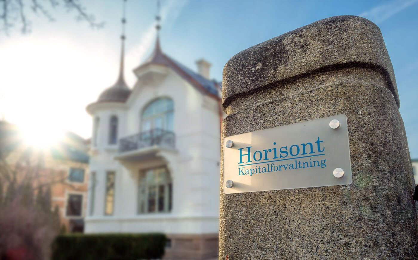 A stone pillar with a sign reading "Horisont Kapitalforvaltning" in front of a white, ornate building with a turret and balconies, under a bright sky with the sun shining.