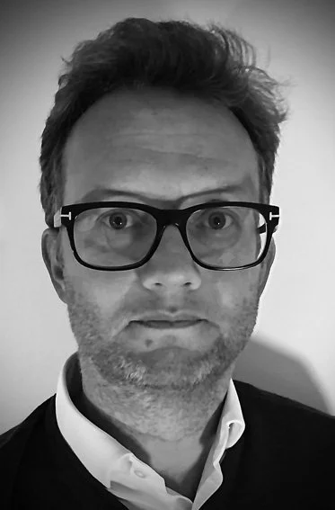 Black and white portrait of a man with glasses, short wavy hair, and a beard, wearing a white shirt and dark jacket.