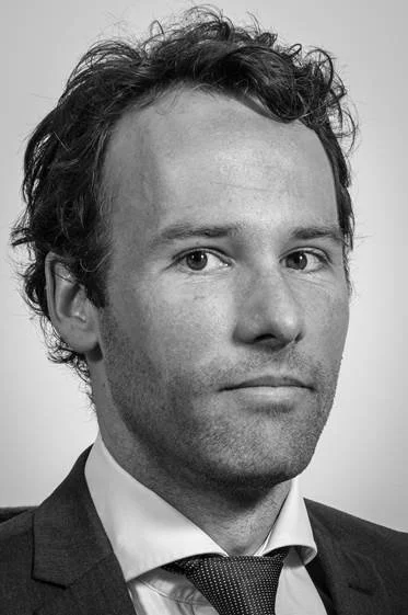 Black and white close-up portrait of a man with curly hair, wearing a suit and tie, looking at the camera with a serious expression.