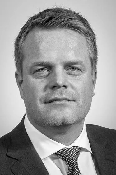 Black and white portrait of a man in a suit and tie, with short hair, looking directly at the camera.